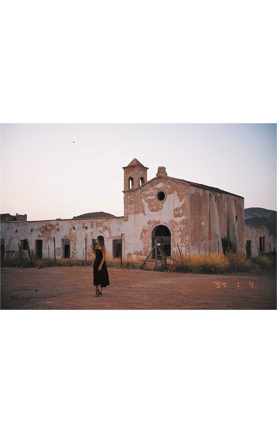 Fotografía de una mujer adulta caminando sola frente a un edificio en ruinas en un entorno rural.