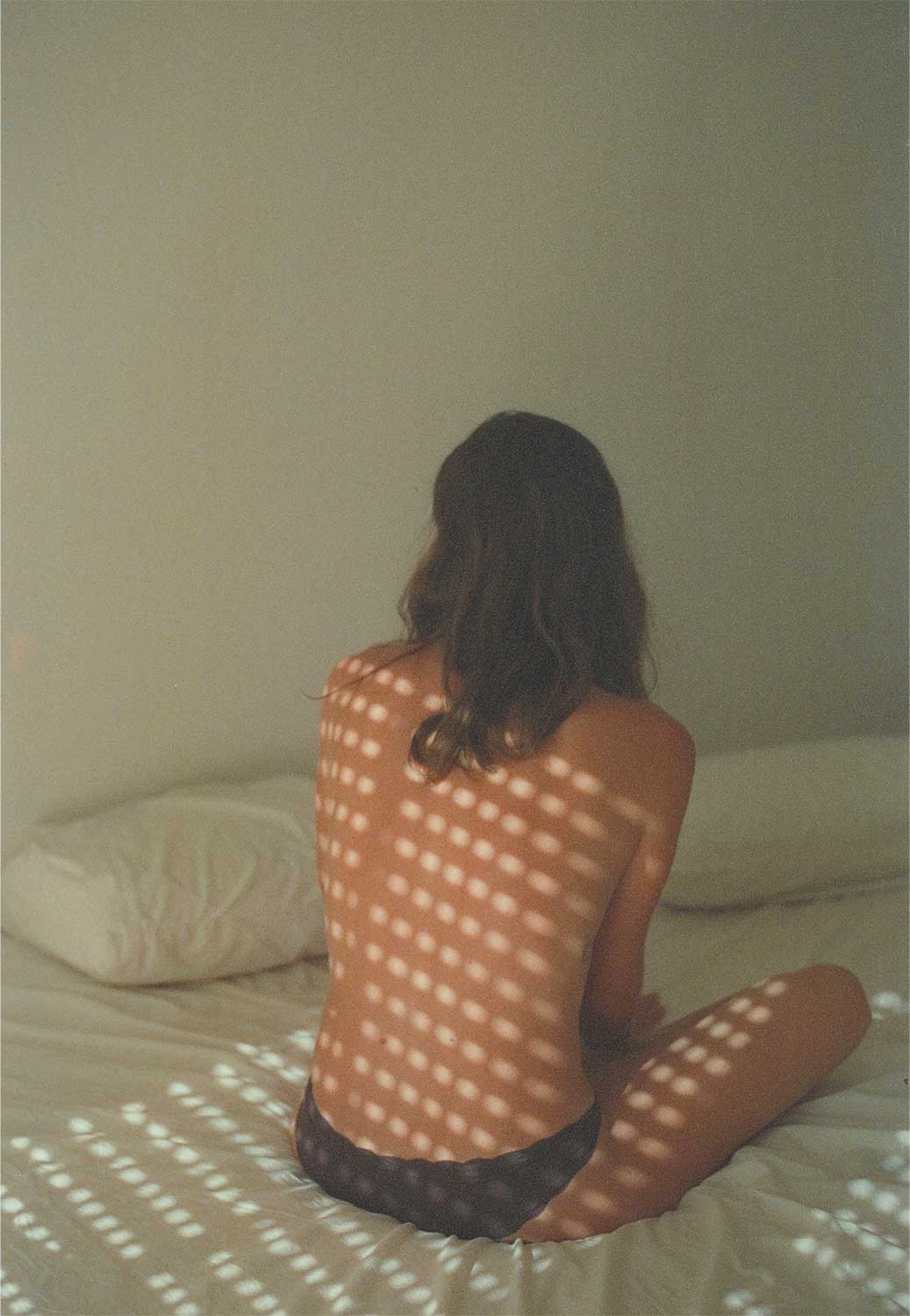 Fotografía de una persona adulta sentada sobre una cama de espaldas, con un patrón de luz proyectado sobre la piel.