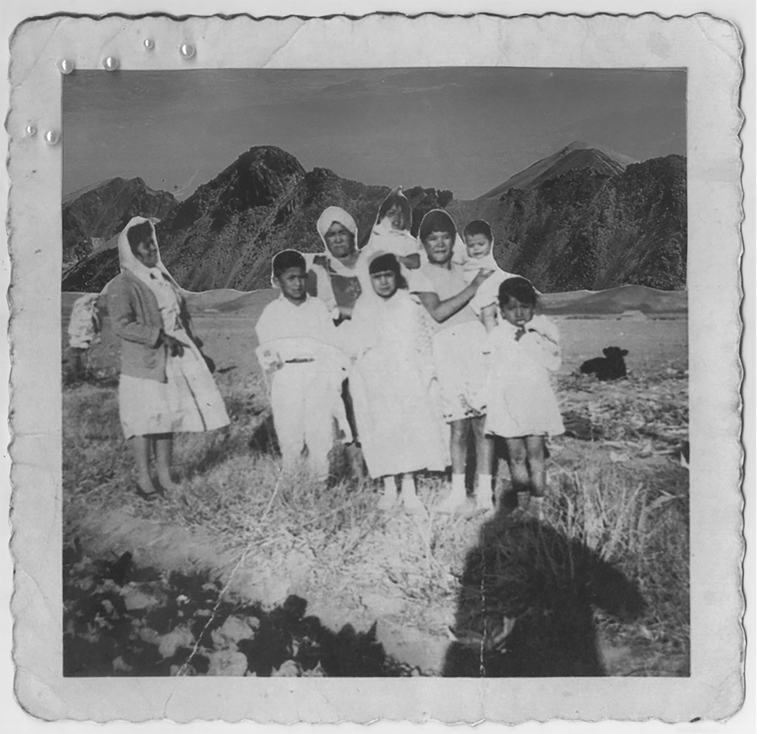 Fotografía en blanco y negro, enmarcada. El marco tiene volantes y algunas perlas pegadas en la parte superior izquierda. En la fotografía aparece una familia indígena con mujeres y niños y niñas en edad temprana, todos vestidos de blanco. A la izquierda, una mujer de mediana edad, también indígena, mira al grupo sonriendo. Se ha recortado en fondo para falsearlo y se ha pegado la fotografía de unas montañas. El recorte es sencillo y se aprecia sin problemas. Al pie de la foto, aparece la alargada ombra del fotógrafo.