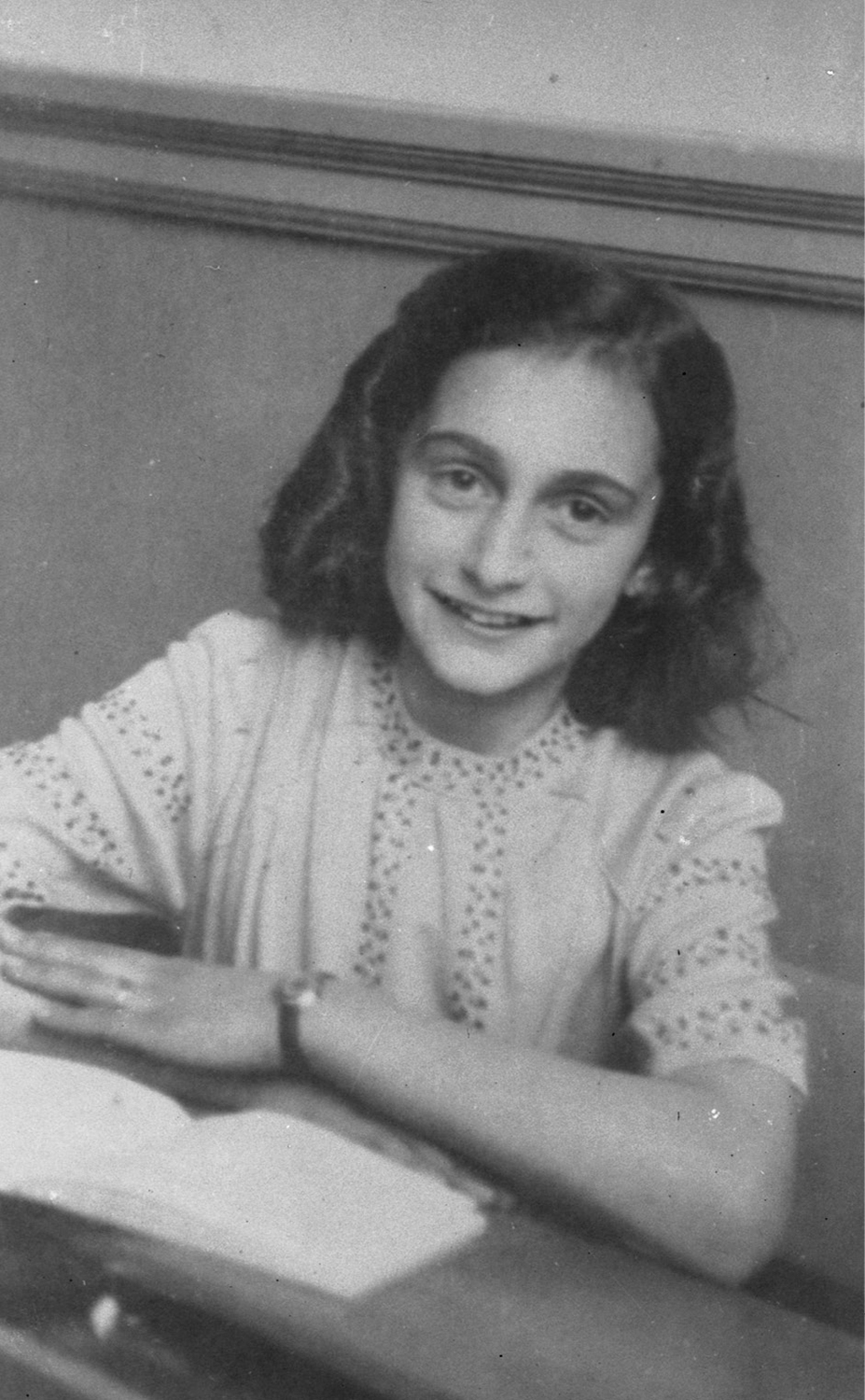 Fotografía en blanco y negro de Anne Frank mirando a cámara sentada en una mesa con un libro abierto delante.