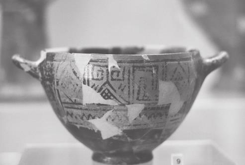 Fotografía en blanco y negro de una copa de cerámica, con dos asas laterales y diversos dubijos geométricos en su superficie, ahora gastada por el paso del tiempo, y con zonas sin pintura, lisas.