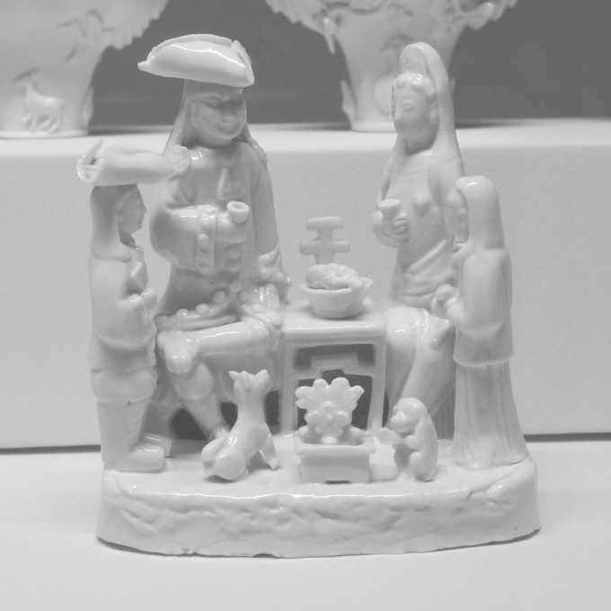 Fotografía de una figura de porcelana blanca que representa una escena doméstica con varias personas reunidas alrededor de una mesa; dos figuras principales permanecen sentadas frente a frente, una de ellas con un sombrero de ala ancha y el brazo extendido hacia la mesa, mientras la otra sostiene un pequeño recipiente; alrededor aparecen cuatro figuras de menor tamaño, dos de pie a ambos lados y dos situadas en la parte frontal junto a la base; sobre la mesa se disponen varios recipientes y alimentos en miniatura, y en primer plano se representa un pequeño brasero o recipiente decorativo.