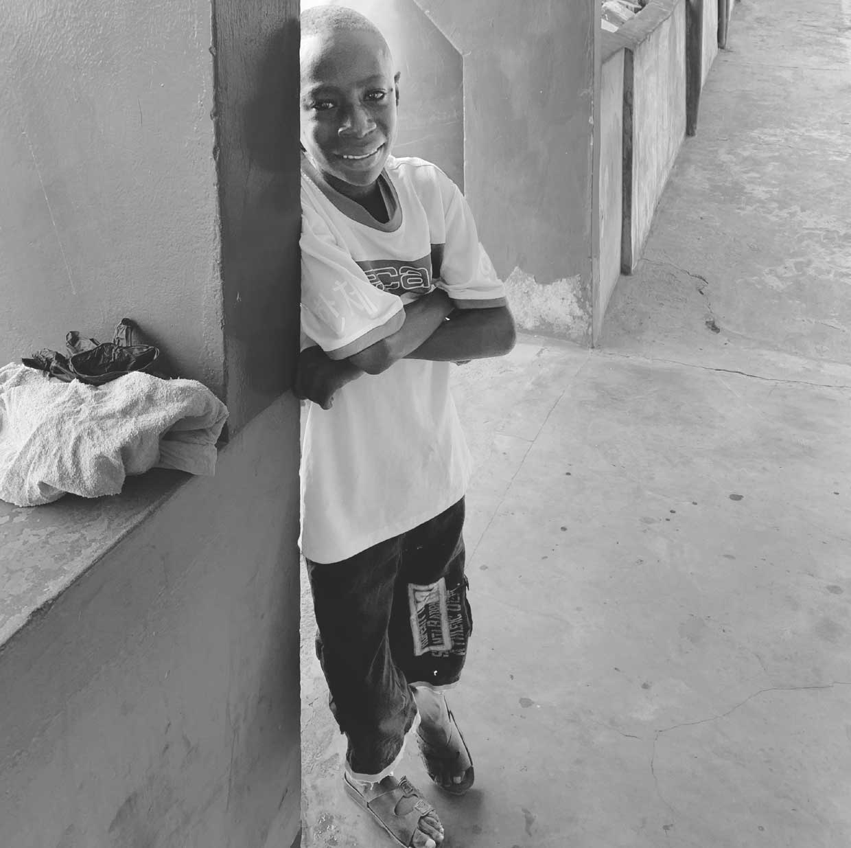 Fotografía documental de Henry de pie en un pasillo, apoyado con la espalda en una pared a la izquierda, brazos cruzados, camiseta de manga corta, pantalón corto y sandalias, una bolsa o bulto sobre un banco de obra en la esquina inferior izquierda, el pasillo se prolonga hacia el fondo a la derecha.