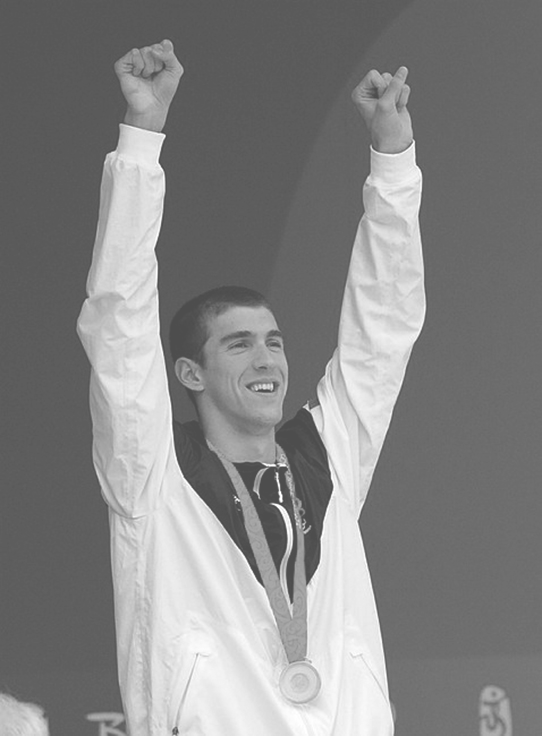 Michael Phelps de pie con ambos brazos extendidos hacia arriba y las manos cerradas, lleva una chaqueta deportiva y una medalla colgada del cuello.